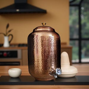 May include: A cylindrical copper water dispenser featuring a hammered texture, a removable lid with a small knob, and a silver-toned metal spigot at the base. The vessel has a warm metallic finish and is positioned upright on a flat surface.