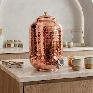 May include: A large, cylindrical, hammered copper water dispenser with a matching lid and a metal spigot at the base. The surface features a textured, dimpled pattern that reflects light, and the dispenser sits on a flat, circular base.