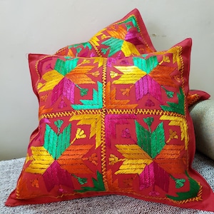 Peut inclure: Deux coussins de canapé rouges avec des motifs géométriques brodés colorés. Les motifs sont réalisés avec du fil vert, jaune, orange et rose.