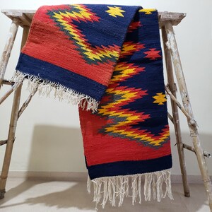 K&ouml;nnte beinhalten: Ein roter, gelber und blauer gewebter Teppich mit geometrischem Muster. Der Teppich hat Fransen an den Enden und ist &uuml;ber einen Holzhocker gelegt.