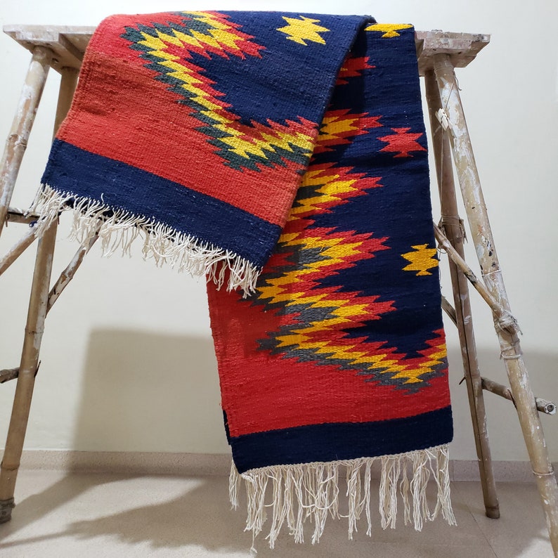 K&ouml;nnte beinhalten: Ein roter, gelber und blauer gewebter Teppich mit geometrischem Muster. Der Teppich hat Fransen an den Enden und ist &uuml;ber einen Holzhocker gelegt.