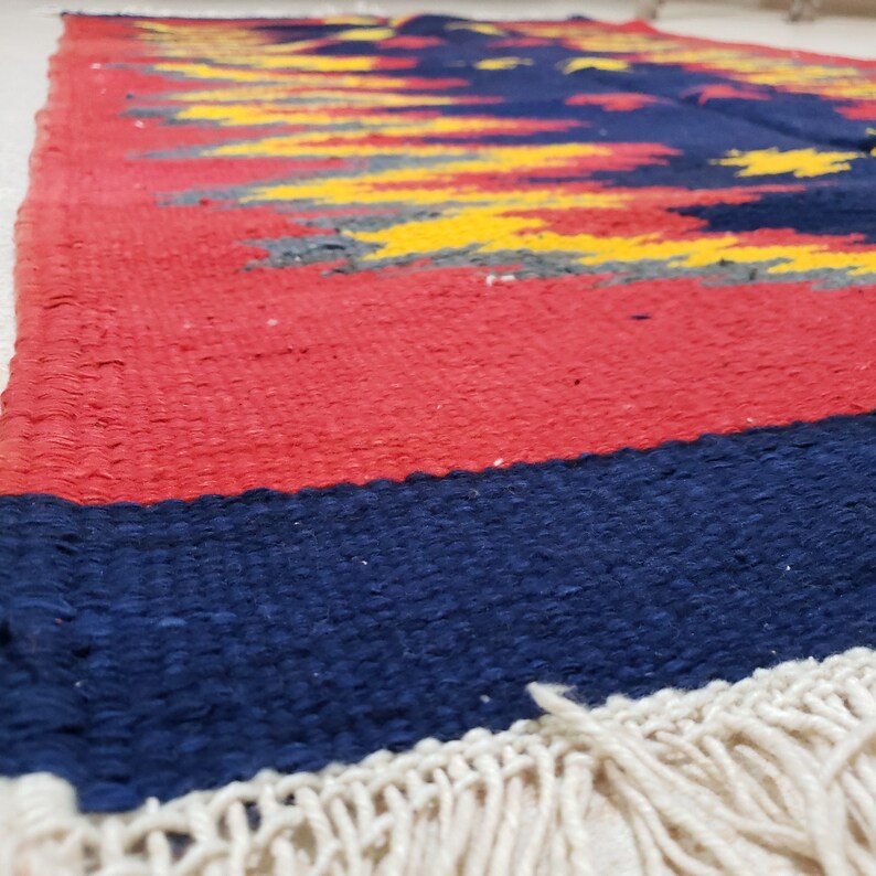 K&ouml;nnte beinhalten: Ein gewebter Teppich mit einem abstrakten Muster in Rot, Gelb, Blau und Grau. Der Teppich hat einen Fransenrand.