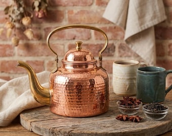 Puede incluir: Una tetera de cobre martillado con un pico curvo y un asa de tono latón. La tetera presenta una tapa redondeada con un pequeño pomo. Junto a ella hay dos tazas de cerámica, una beige y otra azul oscuro, además de dos pequeños cuencos de cristal.