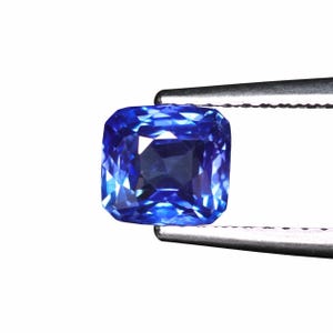 May include: A vibrant, square-cut sapphire gemstone, held by silver tweezers against a white background. The sapphire displays a deep, rich blue hue with multiple facets, reflecting light. The gemstone's shape is a cushion cut.