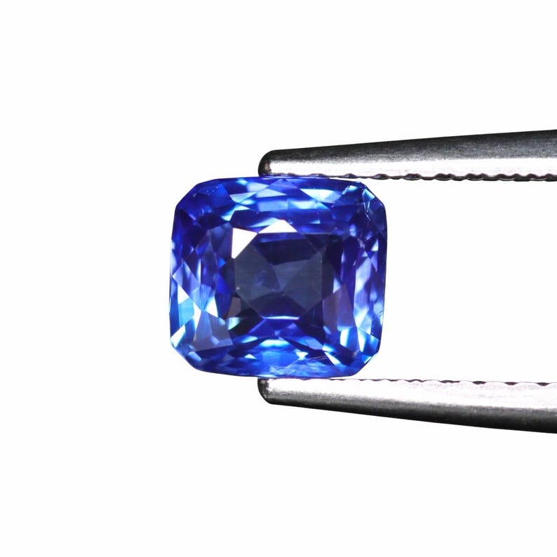 May include: A vibrant, square-cut sapphire gemstone, held by silver tweezers against a white background. The sapphire displays a deep, rich blue hue with multiple facets, reflecting light. The gemstone's shape is a cushion cut.