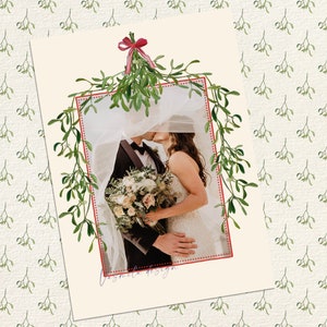 May include: A wedding photo of a couple kissing under a sprig of mistletoe with a red ribbon. The photo is framed with a red dotted line and surrounded by a green mistletoe pattern.