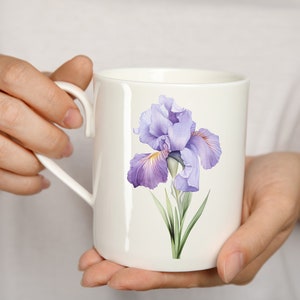 K&ouml;nnte beinhalten: Eine wei&szlig;e Keramiktasse mit einem Design aus einer lila und gelben Iris-Blume.