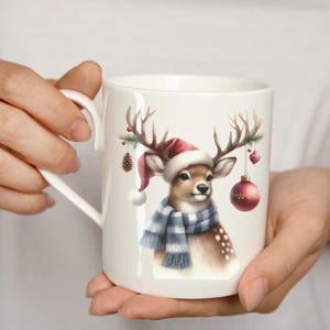 May include: White ceramic mug with a Christmas-themed design featuring a deer wearing a red Santa hat and a blue and white scarf. The deer has antlers with a pine branch and ornaments hanging from them.
