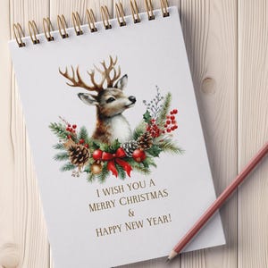May include: A white spiral notebook with a watercolor illustration of a deer with antlers surrounded by a wreath of pine branches, red berries, and a red bow. The text "I wish you a Merry Christmas & Happy New Year!" is printed below the illustration.