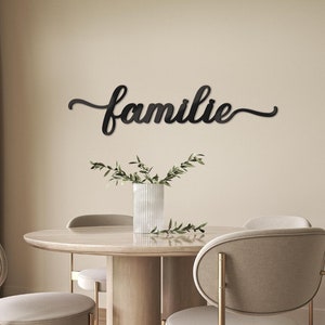 Könnte beinhalten: Ein schwarzes Metall-Wandschild mit dem Wort "familie" in Schreibschrift. Das Schild ist an einer beigefarbenen Wand über einem runden Holztisch mit vier Stühlen angebracht. Auf dem Tisch steht eine weiße Vase mit Grünzeug.