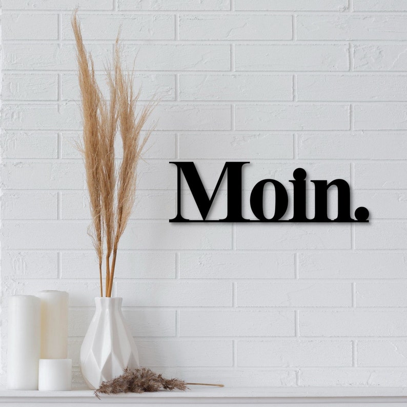 May include: Black metal wall decor with the word "Moin."  The decor is hanging on a white brick wall.