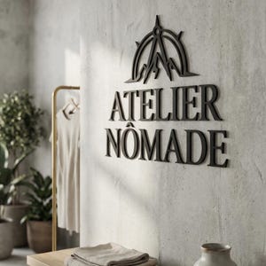 Könnte beinhalten: Schwarze Wandkunst mit dem Schriftzug "ATELIER NOMADE" in fetter Schrift. Darüber ein Kompass- und Bergdesign. Die Kunst ist an einer strukturierten, hellgrauen Wand angebracht, was auf eine minimalistische Ästhetik hindeutet.