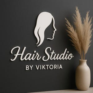 Könnte beinhalten: Weißes Logo für ein Haarstudio mit dem Profil einer Frau und dem Schriftzug "Hair Studio BY VIKTORIA" in eleganter Schrift. Eine Vase mit getrocknetem Pampasgras befindet sich auf der rechten Seite des Bildes, vor einem dunkelgrauen Hintergrund.