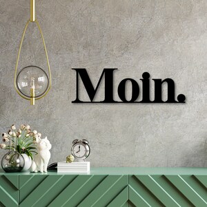 May include: Black metal wall decor with the word "Moin." in bold, block letters. The decor is hanging on a gray concrete wall above a green dresser with a white cat figurine and a clock.