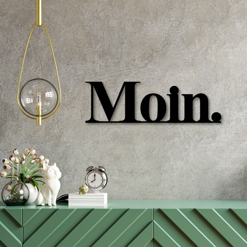 May include: Black metal wall decor with the word "Moin." in bold, block letters. The decor is hanging on a gray concrete wall above a green dresser with a white cat figurine and a clock.