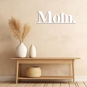 May include: White wooden sign with the word "Moin" on a light beige wall. The sign is hanging above a wooden console table with two white vases and a woven basket on the bottom shelf.