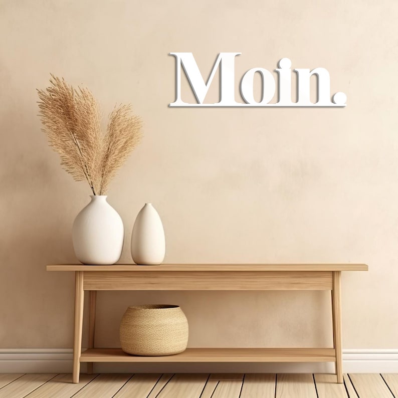 May include: White wooden sign with the word "Moin" on a light beige wall. The sign is hanging above a wooden console table with two white vases and a woven basket on the bottom shelf.