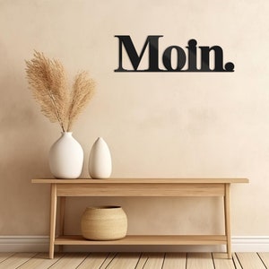 May include: Black wooden wall decor with the word "Moin." displayed on a beige wall above a wooden console table with a woven basket and two white ceramic vases.