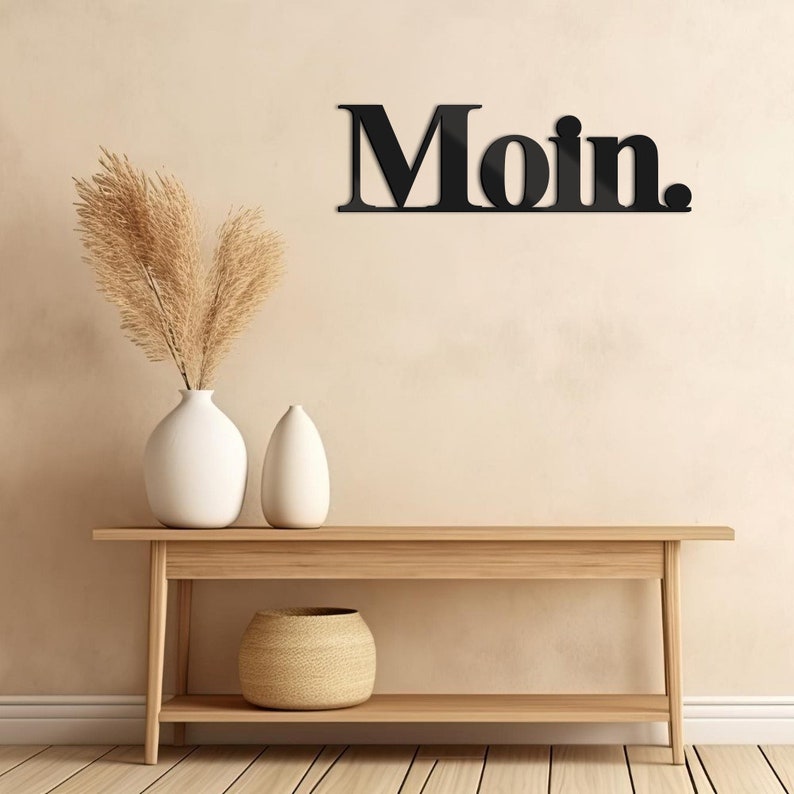 May include: Black wooden wall decor with the word "Moin." displayed on a beige wall above a wooden console table with a woven basket and two white ceramic vases.