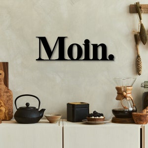May include: Black metal wall decor with the word "Moin." in bold letters. The decor is hanging on a light beige wall. There are various kitchen items on a wooden counter below the decor.