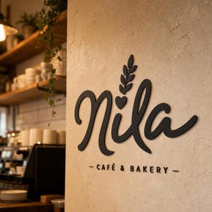 Könnte beinhalten: Ein schwarzes Wand-Schild mit dem Wort Nila in einer geschwungenen Schreibschrift, ergänzt durch ein kleines Herz und einen Blattzweig über dem i. Unter dem Haupttext stehen die Wörter CAFÉ & BAKERY in einer schlichten, serifenlosen Schriftart.