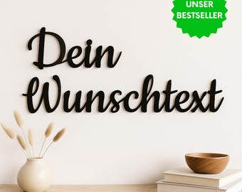 May include: Black wall-mounted lettering in a cursive script font displaying the German text 'Dein Wunschtext'. The letters are arranged in two lines, with 'Dein' on the top and 'Wunschtext' on the bottom, featuring a smooth, matte finish.