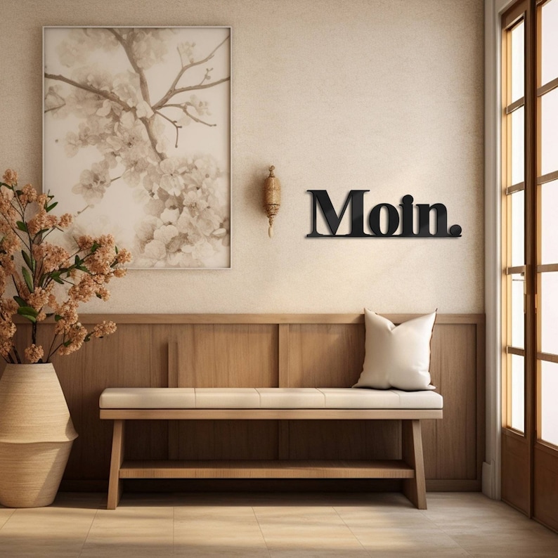 May include: A black metal wall sign with the word "Moin." in bold, block letters. The sign is hanging on a beige wall above a wooden bench with a white cushion. A floral painting hangs on the wall to the left of the sign.