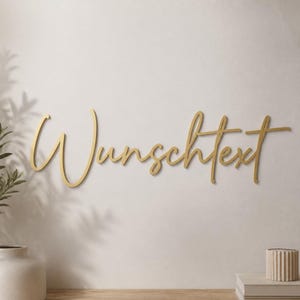 Könnte beinhalten: Ein goldfarbener Wandschriftzug mit dem Wort "Wunschtext" in einer durchgehenden, fließenden Schreibschrift. Die Buchstaben sind miteinander verbunden, haben eine glatte, metallische Oberfläche und sind horizontal an einer hellen Wand angebracht.