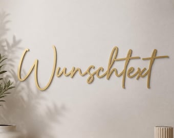 May include: A gold-colored wall sign featuring the word "Wunschtext" written in a continuous, flowing cursive script. The letters are connected and have a smooth, metallic finish, mounted horizontally against a plain, light-colored wall surface.