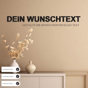 Könnte beinhalten: Ein schwarzes Holzschild mit dem Text "DEIN WUNSCHTEXT" und dem Satz "GESTALTE DIR DEINEN INDIVIDUELLEN TEXT". Das Schild ist an einer beigefarbenen Wand montiert. Auf einer weißen Kommode steht eine Vase mit Trockenblumen und eine kleinere Vase.