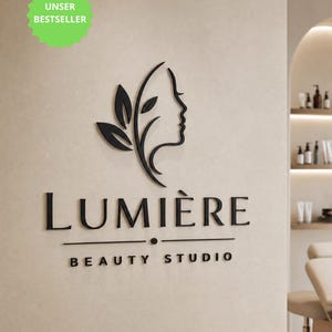 Könnte beinhalten: Ein schwarzes 3D-Wandschild mit einem Gesichtsprofil kombiniert mit Blattformen, dem Text "LUMIÈRE" in großen Buchstaben und dem Text "BEAUTY STUDIO" unter einer horizontalen Linie mit einem mittigen Punkt, montiert an einer hellbeigen Wand.