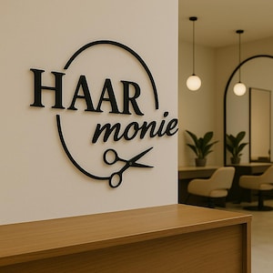 Könnte beinhalten: Schwarzes Schild mit der Aufschrift "HAAR monie" und einer Schere in einem Kreis. Das Schild ist an einer weißen Wand angebracht. Im Hintergrund ist das Innere eines Friseursalons zu sehen.
