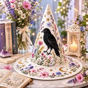 May include: A white conical hat featuring an embroidered black crow surrounded by colorful wildflowers, lavender, and gold celestial stars and moons. The fabric has a quilted texture, and the design covers both the crown and the wide brim.