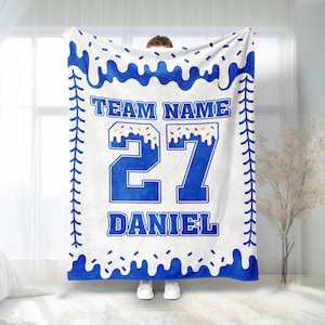 May include: A white blanket with blue accents and text. The blanket features a blue border with a dripping design, a baseball stitch pattern, and the words "TEAM NAME", "27", and "DANIEL" in blue. The number 27 has a colorful sprinkle design.