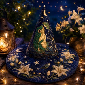 May include: A dark blue quilted pointed hat featuring an embroidered green frog, a moth, and white lily flowers. The design includes gold stitched stars and crescent moons across the fabric, creating a celestial pattern on the conical shape.
