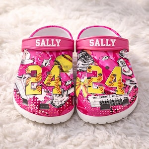 May include: A pair of pink clogs with baseball-themed graphics. The clogs feature the name "SALLY" on the strap, the number "24", and baseball equipment illustrations. The clogs have a white sole.