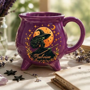 May include: A purple cauldron-shaped ceramic mug with three small feet and a handle. The front features a graphic of a witch in a pointed hat riding a broomstick against a large orange moon, surrounded by stars and the name Sabrina in script.