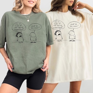May include: Two t-shirts, one sage green and one cream, each featuring a black and white cartoon. The cartoon shows two figures with speech bubbles. The text in the speech bubbles reads "I'm so sick of not being understood!" and "What do you mean?"