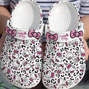 May include: White clogs featuring a repeating pattern of Hello Kitty characters, bows, and speech bubbles. The clogs have a pink glitter bow on the strap and the words "Hello Kitty" printed on the side. The design includes black leopard print spots.
