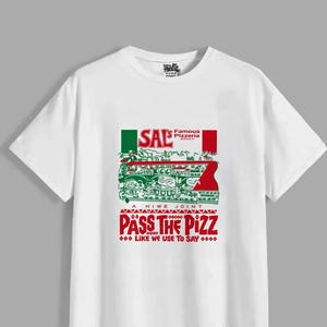 Puede incluir: Una camiseta blanca de manga corta con un estampado gr&aacute;fico que incluye el texto Sal's Famous Pizzeria Brooklyn, A Hiwe Joint y Pass The Pizz Like We Use To Say. El dise&ntilde;o presenta detalles en verde y rojo y una ilustraci&oacute;n.