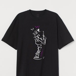 Może przedstawiać: Czarny t-shirt z krótkim rękawem z białym i fioletowym nadrukiem graficznym. Grafika przedstawia saksofon, koronę i stylizowany tekst. T-shirt jest wykonany z miękkiego materiału.
