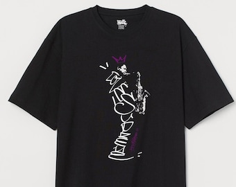May include: A black short-sleeved t-shirt featuring a white graphic print of a person playing a saxophone with a purple crown above the head. The design includes stylised white text and a small purple signature near the bottom of the graphic.