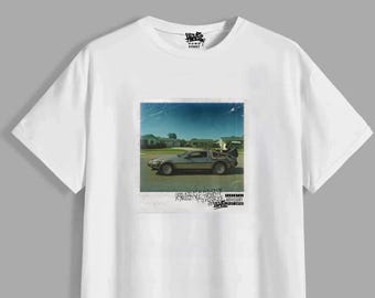 May include: A white short-sleeved t-shirt featuring a central graphic print of a silver sports car parked on a suburban street. The graphic resembles a polaroid photo with text below it, including the words Good Kid, A Freestyle Tribute, and Parental Advisory.