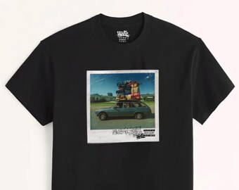 May include: A black short-sleeve t-shirt featuring a central graphic of a polaroid-style photograph. The image depicts a teal station wagon heavily loaded with luggage on its roof, with text below reading Good Kid M.A.A.D City.