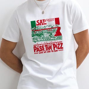 Puede incluir: Una camiseta blanca de manga corta con un estampado gr&aacute;fico en rojo, blanco y verde. El dise&ntilde;o incluye el texto SAL'S FAMOUS PIZZERIA, A HIWE JOINT, PASS THE PIZZ y LIKE WE USE TO SAY, junto a una ilustraci&oacute;n de un edificio.