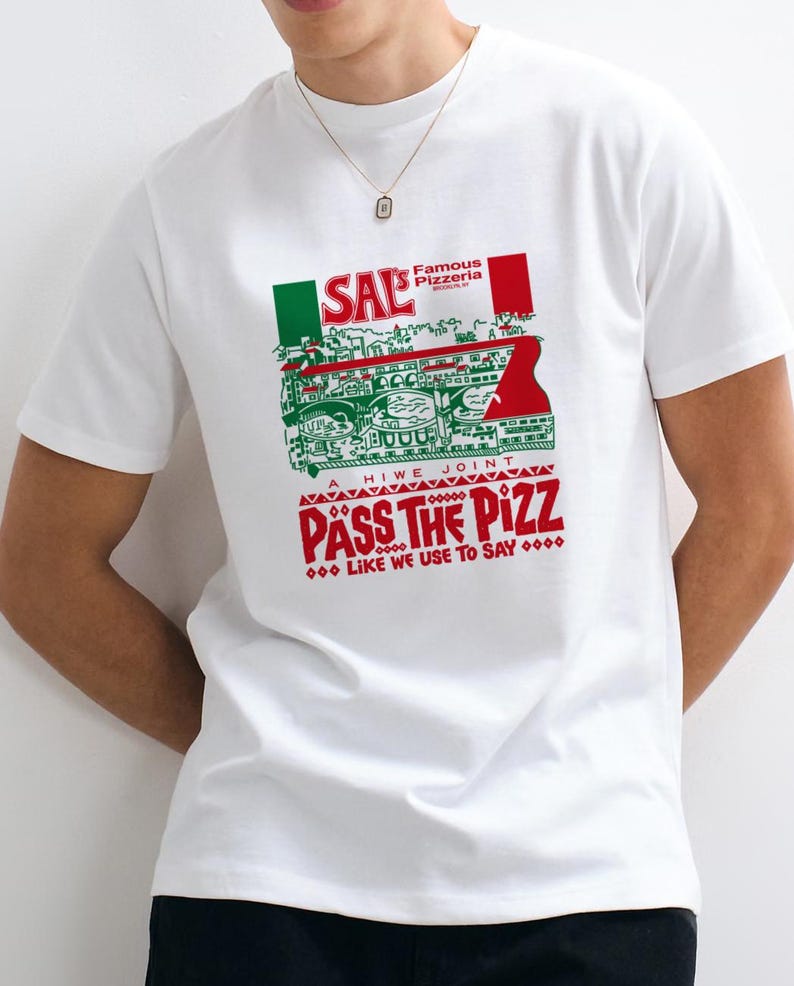 Puede incluir: Una camiseta blanca de manga corta con un estampado gr&aacute;fico en rojo, blanco y verde. El dise&ntilde;o incluye el texto SAL'S FAMOUS PIZZERIA, A HIWE JOINT, PASS THE PIZZ y LIKE WE USE TO SAY, junto a una ilustraci&oacute;n de un edificio.