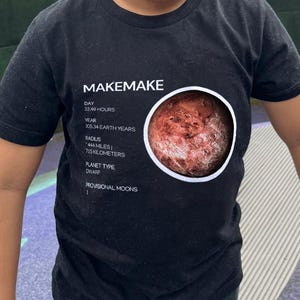 Puede incluir: Un niño lleva una camiseta negra con la palabra "MAKEMAKE" en la parte superior. Debajo, un gráfico de un planeta e información. El niño sonríe. La camiseta muestra datos sobre el planeta.