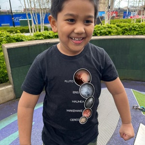 May include: A young boy wearing a black t-shirt with a graphic of the dwarf planets Pluto, Eris, Haumea, Makemake, and Ceres. The graphic is a vertical line of circles with the names of the dwarf planets written next to them. The boy is smiling and looking to the right. He is wearing camouflage pants.