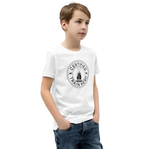 May include: White t-shirt with a black circular graphic that reads "Certified Train Nerd" around a train illustration. The shirt is worn by a person in blue jeans.