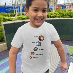 Puede incluir: Un niño pequeño con una camiseta blanca con un gráfico de los planetas enanos Plutón, Eris, Haumea, Makemake y Ceres. El gráfico es en negro, blanco y tonos de marrón y rojo.
